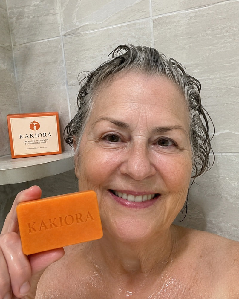 Older woman in the shower holding a KAKIORA soap bar with the box on the shelf